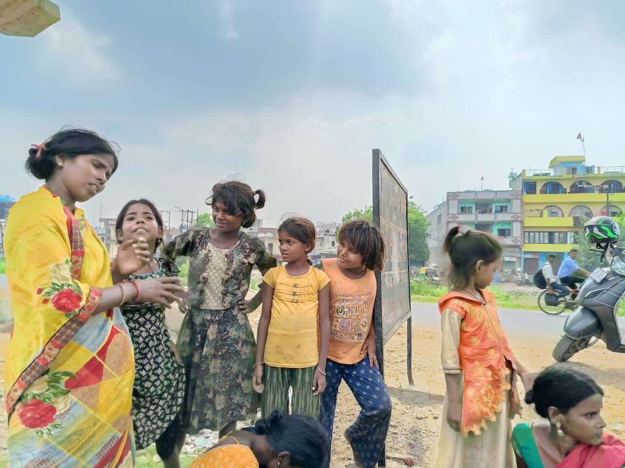 Group of children standing outdoors by a road, with buildings and a passing motorcycle in the background.