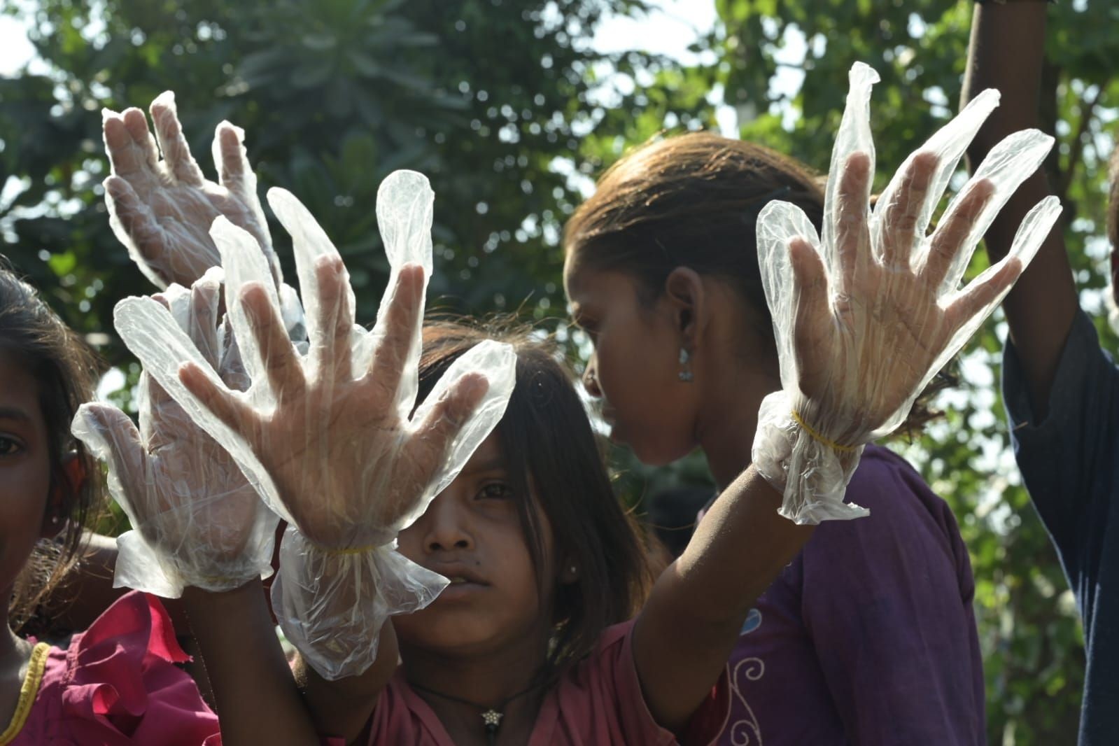 Children wearing plastic gloves, hands raised, outdoors with trees in the background.